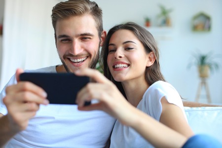 Young couple watching online content in a smart phone sitting on a sofa at home in the living room.の写真素材