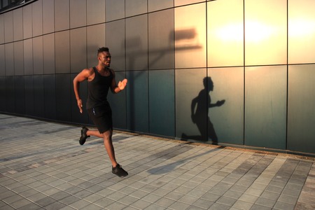 Full length of young African man in sports clothing jogging while exercising outdoors, at sunset or...