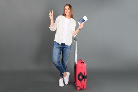 Young tourist girl in summer casual clothes, with red suitcase, passport, tickets isolated on grey...