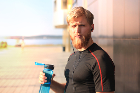 Handsome adult man drinking water from fitness bottle while standing outside, at sunset or sunrise. Runner.の写真素材