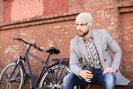Handsome young man in grey coat and hat sitting on a bench relaxed drinking coffee and thinking near his bicycleの写真素材