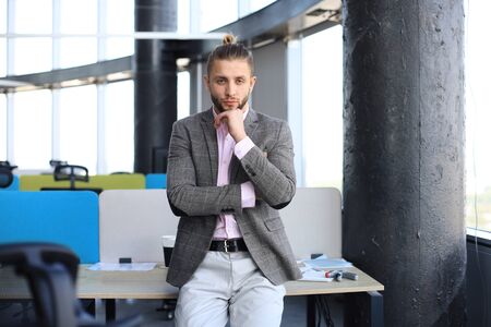 Thoughtful young businessman in casual clothes looking at camera and keeping hand on chin while leaning on the deskの写真素材
