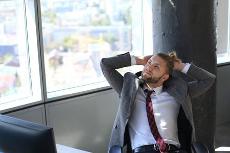Young modern businessman keeping hands behind head and smiling while sitting in the officeの写真素材
