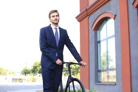 Young stylish business man dressed in suit walking with a bicycle on a city street.の写真素材
