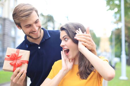Astonished excited couple in summer clothes smiling and holding present box together while standing on city streetの写真素材
