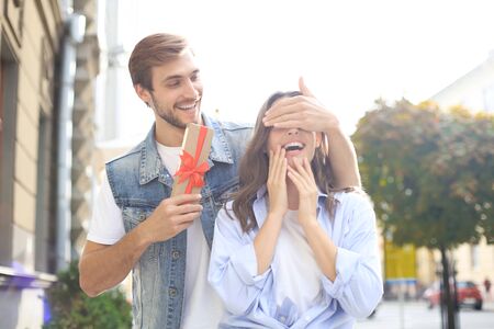 Portrait of a happy young couple hugging while standing together with a present box, man covers womans eyes.の写真素材
