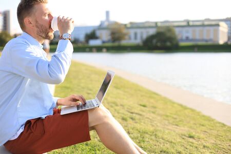Handsome man spending time outdoors at the city, using laptop computer while sitting on a bench and drinking coffee.の写真素材