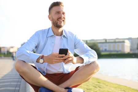 Happy young handsome man sitting on the bench outdoors and using smartphone.の写真素材