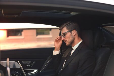 Thoughtful confident businessman keeping hand on glasses while sitting in the luxe car