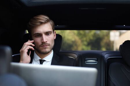 Handsome confident businessman in suit talking on smart phone and working using laptop while sitting...