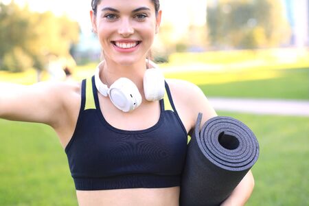 Beautiful happy young sports woman holding fitness rug in urban parkの写真素材