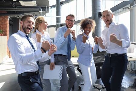 Five happy modern business people are keeping arms raised and expressing joyful while standing in...