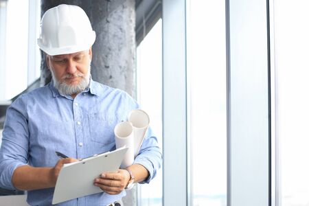 Confident mature business man in hardhat holding blueprint while standing indoors
