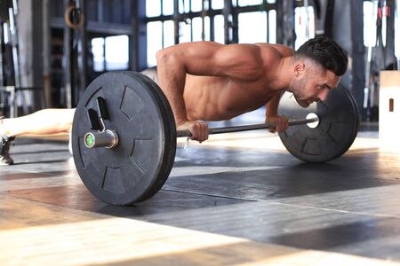 Fit and muscular man doing horizontal push-ups with barbell in gym