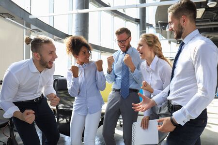 Five happy modern business people are keeping arms raised and expressing joyful while standing in...