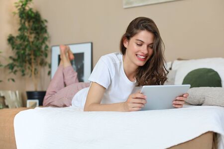Smiling woman is liying on the bed and use her digital tablet