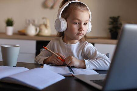 Smiling little girl in headphones handwrite study online using laptop at home, cute happy small...
