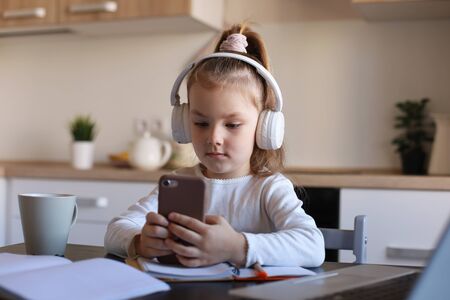 Smart little girl in headphones do homework online class on smartphone, small child in earphones study on Internet, have web conference or lesson during quarantineの写真素材