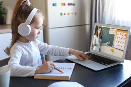 Smart small preschooler girl in headphones watch online lesson and communicate with teacher at home,...