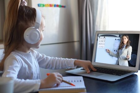 Smart small preschooler girl in headphones watch online lesson and communicate with teacher at home,...