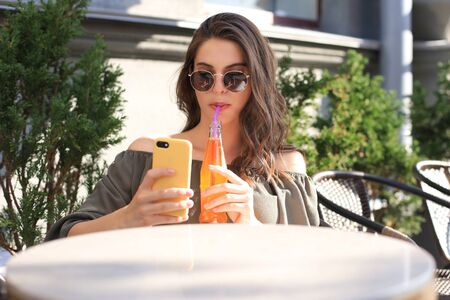 Smiling beautiful brunette woman sitting in street summer cafe, take selfie by mobile phone, drinking juiceの写真素材