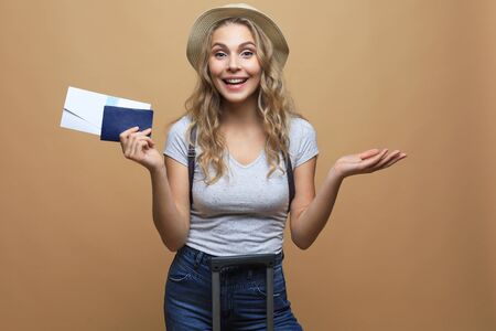 Cheerful blonde woman in summer clothes posing with baggage and holding passport with tickets over beige backgroundの写真素材