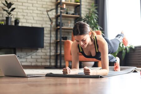 Beautiful slim fitness woman doing a plank exercise watching online tutorials on laptop, training in living room. Healthy lifestyle. Girl goes in for sports at home.の写真素材