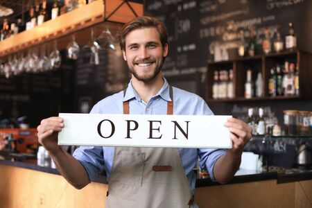 Smiling male owner of coffee shop is standing indoors. He is holding placard and inviting everybody...