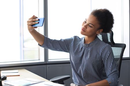 Young african  woman doing selfie for social nets while sitting in creative officeの写真素材