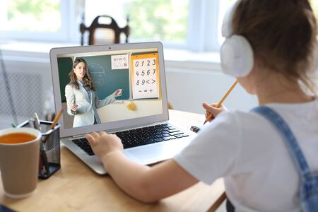 Distance learning. Cheerful little girl girl in headphones using laptop studying through online...