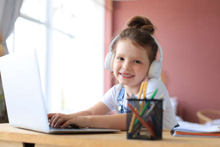 Smiling little girl in headphones handwrite study online using laptop at home, cute happy small...