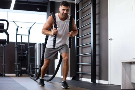 Athletic young man with battle rope doing exercise in functional training fitness gym