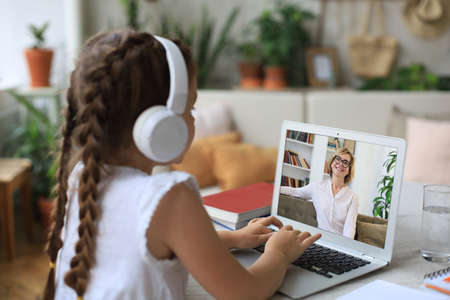 Distance learning. Cheerful little girl girl in headphones using laptop studying through online...