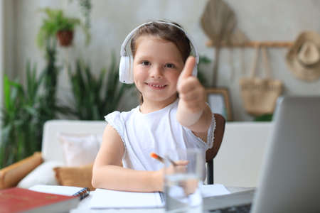 Happy little girl pupil study online using laptop at home, smiling small child show thumb up...