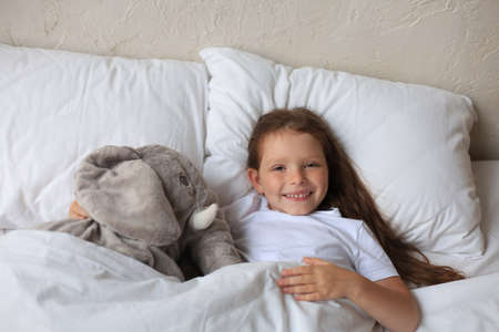 Cute child little girl wake up and lies in the bed with a toy elephantの写真素材