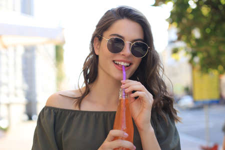 Attractive young woman in sunglasses holds a bottle with juice, looking at camera, outdoors