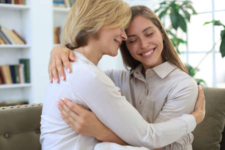 Beautiful middle aged mom and her adult daughter are hugging and smiling while sitting on couch at...
