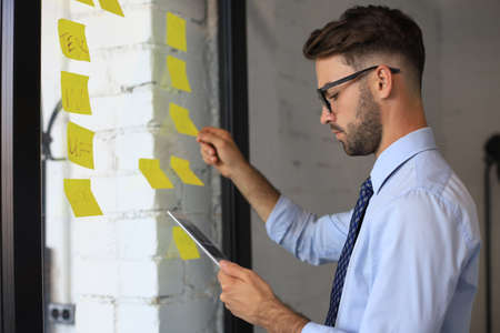 Young modern business man using adhesive notes while standing behind the glass wall in the officeの写真素材