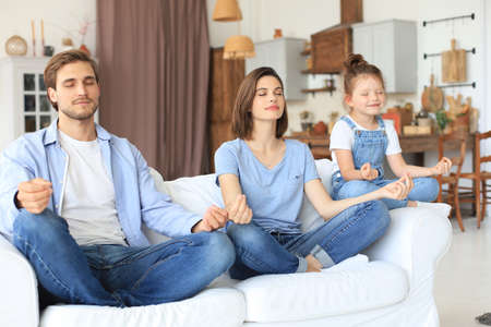 Calm young family with little daughter sit on couch practice yoga together, happy parents with small preschooler girl child rest on sofa meditate relieve negative emotions on weekend at homeの写真素材