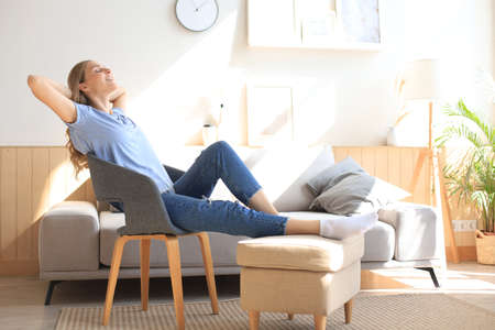 Young woman at home sitting on modern chair in front of window, relaxing in her living room