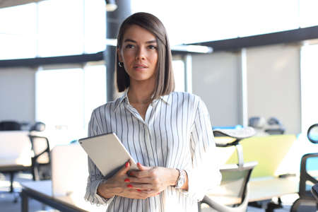 Portrait of young businesswoman holding touch pad while standing in modern office space interior