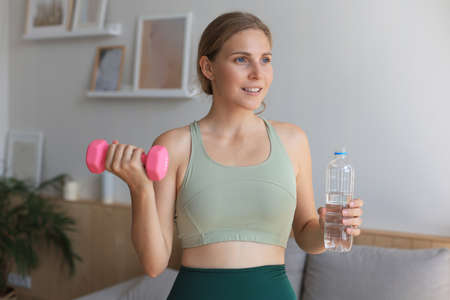 Young beautiful woman standing with dumbbell and drink water after exercising. Attractive female...