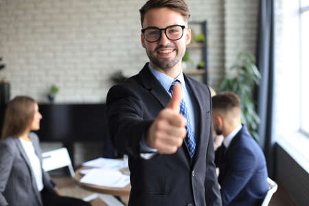 Happy businessman showing his thumb up with colleagues in the background in office