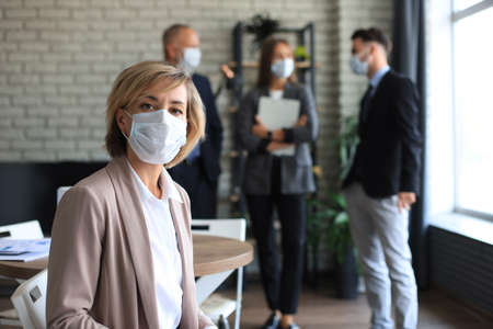 Businesswoman in medical mask with her staff, people group in background at modern bright office...