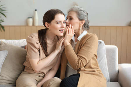 Happy middle mother whispering secret to her smiling daughter at home, gossiping, sharing secret....
