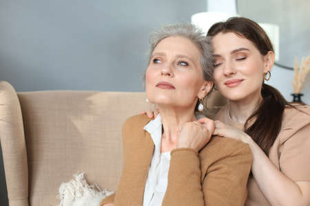 Happy daughter hugging older mother, standing behind chair in living room, enjoying tender moment at homeの写真素材
