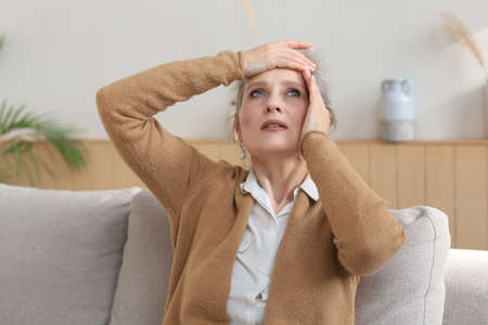 Stressed middle aged woman sit on sofa in living room, lost in thoughtの写真素材