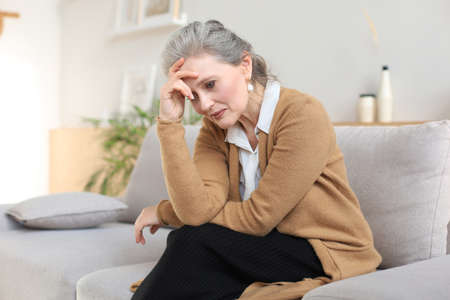 Stressed middle aged woman sit on sofa in living room, lost in thought