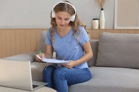 Smiling girl sit near couch watching webinar on laptop. Happy young woman study on online distant...