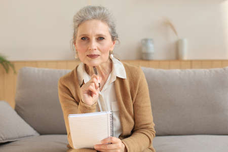 Attractive mature woman sitting on the couch and making notes in diaryの写真素材